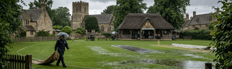 Man pulling cricket pitch cover on rain-soaked cricket ground in village